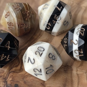 May include: Five multi-sided dice in various colours, including black, white, and brown, with etched numbers. The dice are arranged on a wooden surface, showcasing their geometric shapes and detailed numbering for tabletop gaming.