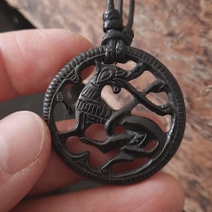 May include: Black circular pendant featuring a carved design of a deer-like creature and a smaller animal, suspended from a black cord. The intricate design highlights the animals' forms, creating a detailed piece of jewellery.