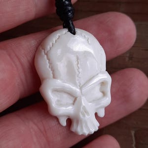 Könnte beinhalten: Weißer, geschnitzter Totenkopf-Anhänger an einer schwarzen Schnur.