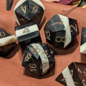 May include: A collection of black and white polyhedral dice with gold numbering. The dice are in various shapes, including a d4, d6, d8, d10, d12, and d20. The dice are made of a glossy material and are arranged on a brown surface.