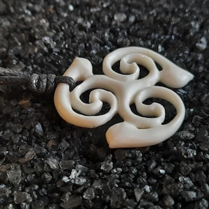 May include: Cream-coloured pendant with a swirling, abstract design, suspended from a black cord. The pendant's smooth surface contrasts with the dark, textured background. A unique, handcrafted piece.