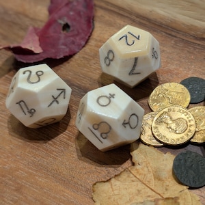 May include: Three ivory-coloured, multi-sided dice with black symbols and numbers, alongside gold coins and other small objects. The dice feature astrological symbols. The items are arranged on a wooden surface with a leaf.