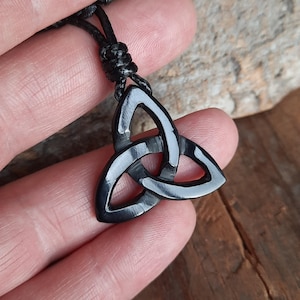 May include: A black Triquetra pendant necklace. The pendant features a three-pointed design, symbolizing interconnectedness. The necklace has a black cord. The pendant is held in a hand.