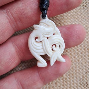 May include: Ivory-colored pendant necklace with a carved dragon design. The pendant is suspended from a black cord. The dragon is depicted in a stylized, swirling form, with intricate details. The necklace is held in a hand.