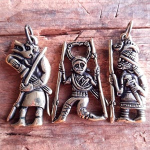 Könnte beinhalten: Drei antike Messinganhänger mit aufwendigen Designs. Die Anhänger zeigen stilisierte Figuren, darunter einen Krieger mit einer Waffe, eine Figur, die Stangen hält, und eine Person mit Kopfschmuck. Die Anhänger sind an kleinen Schlaufen aufgehängt.
