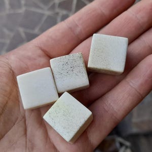 May include: A hand holds five small, off-white square tiles with rounded edges. The tiles have a textured surface, with some showing darker spots. These tiles could be for a game or a craft project.
