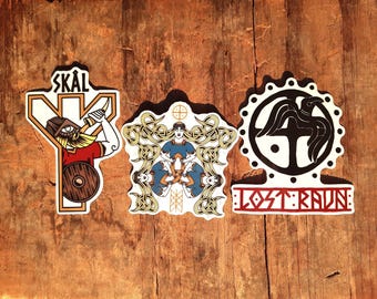 May include: Three stickers with a Viking theme. The first sticker is a white sticker with a black and red illustration of a Viking holding a sword and shield. The sticker has the word "SKÅL" at the top. The second sticker is a white sticker with a black and gold illustration of a woman with long hair. The third sticker is a white sticker with a black illustration of a raven with a cross in the center. The sticker has the text "LOST:RAUN" at the bottom.