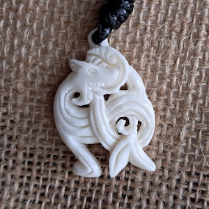 May include: Cream-coloured pendant necklace featuring a carved design of a stylised animal with swirling patterns. The pendant is suspended from a black cord. The background is a textured, neutral-toned fabric.