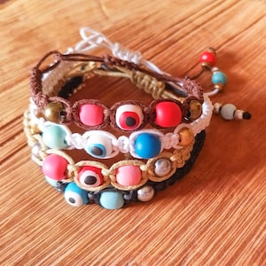 Puede incluir: Cuatro pulseras superpuestas con cuentas de ojo turco en azul, rojo y blanco. Las pulseras están hechas con cordones marrones, blancos, negros y dorados y presentan cuentas de varios colores.