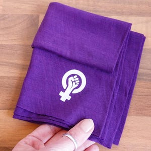 Puede incluir: Paño morado doblado con un gráfico blanco de un puño levantado dentro de un círculo y una cruz debajo. El paño está cuidadosamente apilado, con el gráfico visible en la capa superior. El fondo es una superficie de madera marrón claro.