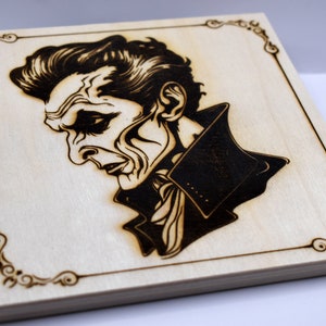 May include: A wood-burned portrait of a man with a dark, slicked-back hairstyle, wearing a dark jacket with a white scarf. The portrait is framed with a decorative border.