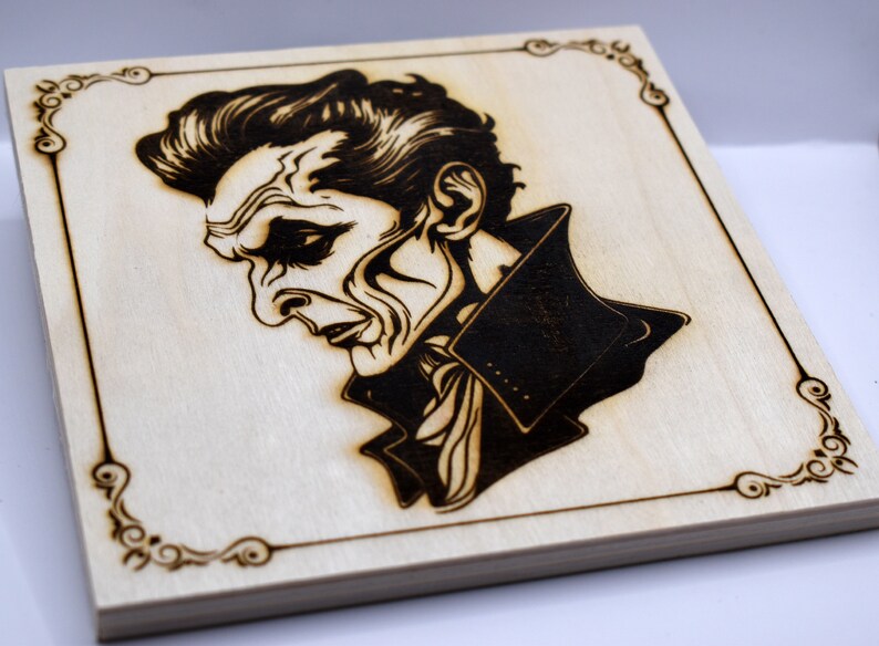 May include: A wood-burned portrait of a man with a dark, slicked-back hairstyle, wearing a dark jacket with a white scarf. The portrait is framed with a decorative border.