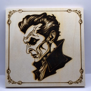 May include: A wood-burned image of a man with a dark, serious expression. He is wearing a dark jacket with a high collar. The image is framed with a decorative border.