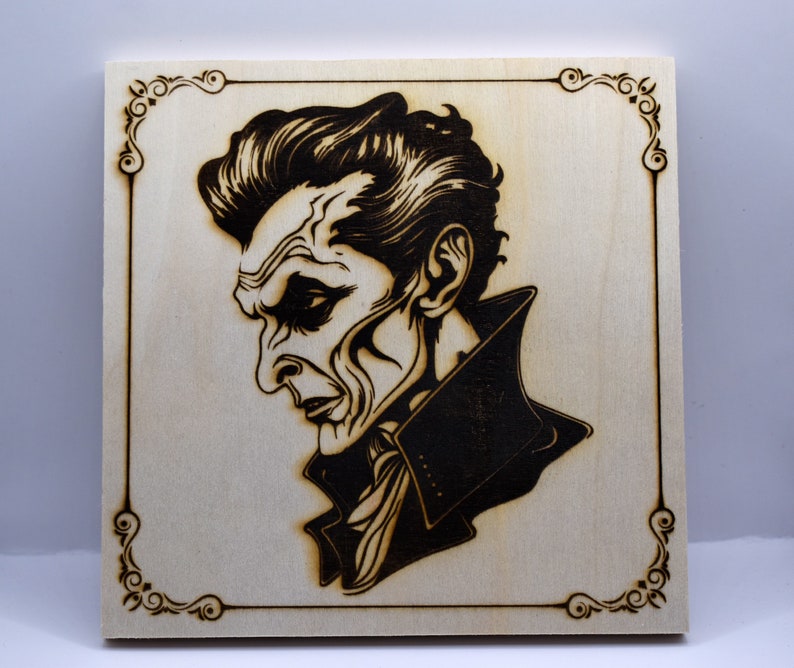 May include: A wood-burned image of a man with a dark, serious expression. He is wearing a dark jacket with a high collar. The image is framed with a decorative border.