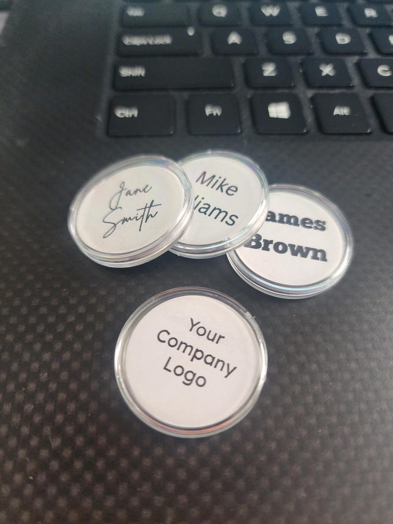 K&ouml;nnte beinhalten: Eine Sammlung klarer, runder Namensschilder mit wei&szlig;en Eins&auml;tzen. Jedes Schild zeigt einen Namen oder Text, darunter "Jane Smith", "Mike Viams", "James Brown" und "Your Company Logo". Die Schilder liegen auf einer dunklen Oberfl&auml;che.