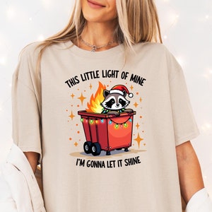 May include: A beige t-shirt with a graphic of a raccoon wearing a Santa hat and sitting in a red trash can with Christmas lights. The text on the shirt reads "This little light of mine I'm gonna let it shine."