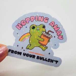 May include: A holographic sticker with a cartoon frog holding a cup of orange juice. The frog is running and the text says "Hopping Away From Your Bullsh*t".