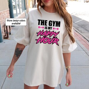 Peut inclure: Un t-shirt blanc surdimensionn&eacute; avec le texte "The Gym is my Happy Hour" en lettres noires et roses. Le texte est dans une police de caract&egrave;res graphique et audacieuse.