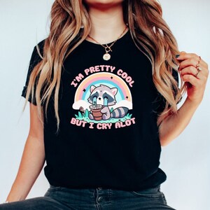 May include: Black t-shirt with a graphic of a crying raccoon holding a cup of coffee. The text on the shirt reads "I'm pretty cool but I cry alot" with a rainbow behind the raccoon.