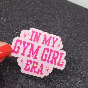 以下が含まれることがあります： ピンクのキラキラしたステッカーに、ピンクの文字で「In My Gym Girl Era」と書かれています。