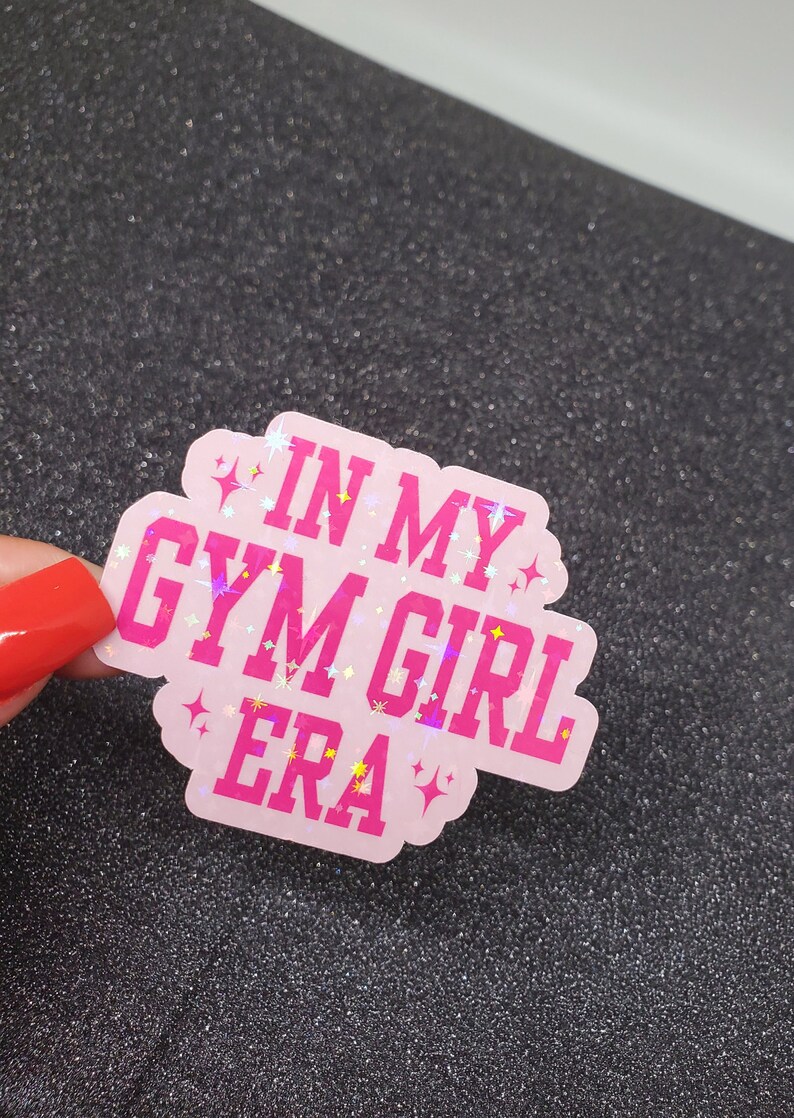 以下が含まれることがあります： ピンクのキラキラしたステッカーに、ピンクの文字で「In My Gym Girl Era」と書かれています。
