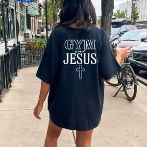 May include: A black t-shirt with white text that reads "GYM and JESUS" with a cross underneath. The person wearing the shirt is standing on a sidewalk in front of a building.