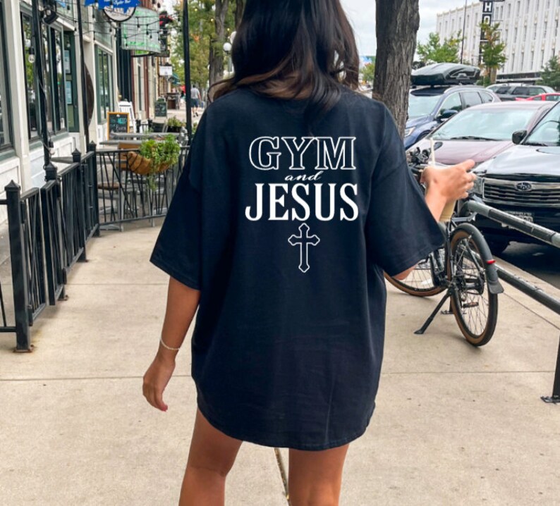 May include: A black t-shirt with white text that reads "GYM and JESUS" with a cross underneath. The person wearing the shirt is standing on a sidewalk in front of a building.