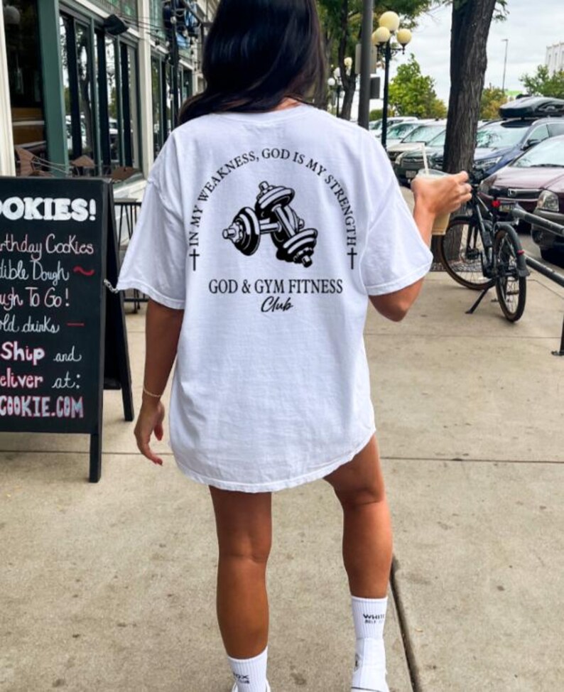 May include: White t-shirt with a black graphic of dumbbells and the text "In my weakness, God is my strength + God & Gym Fitness Club".