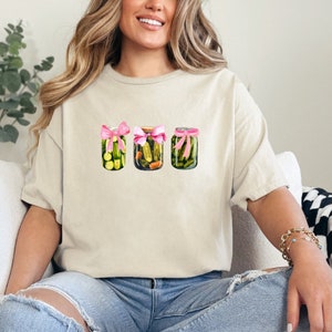 Pode incluir: Uma camiseta branca com um gr&aacute;fico de tr&ecirc;s frascos de vidro cheios de picles verdes, cada um amarrado com uma fita rosa.
