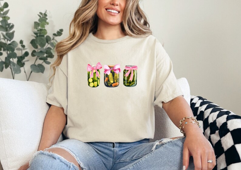 Pode incluir: Uma camiseta branca com um gr&aacute;fico de tr&ecirc;s frascos de vidro cheios de picles verdes, cada um amarrado com uma fita rosa.