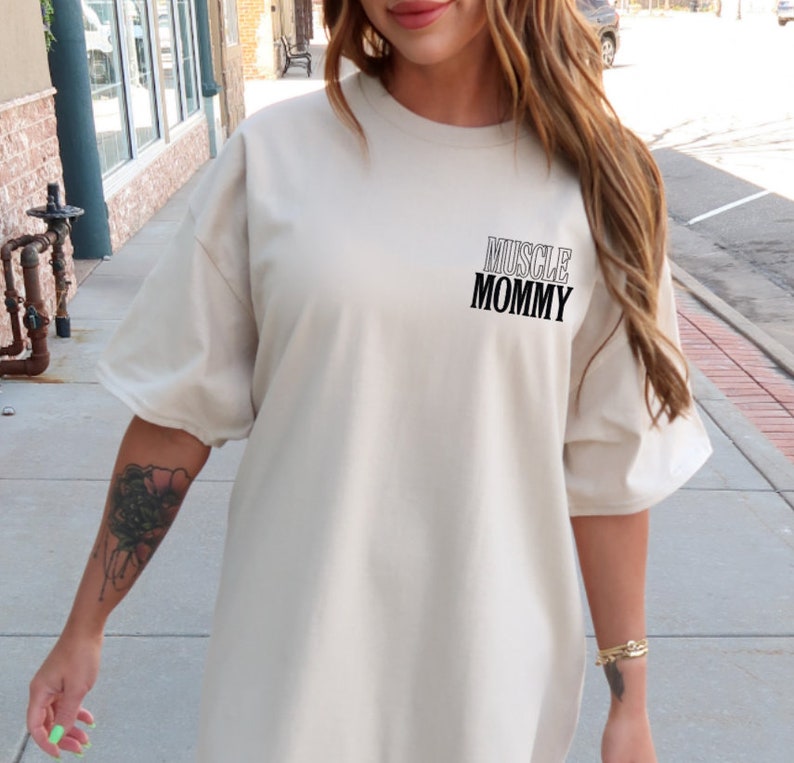 May include: A light beige oversized t-shirt with the text "MUSCLE MOMMY" printed on the front.