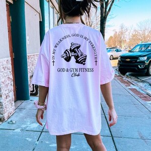 May include: Pink t-shirt with a black graphic of dumbbells and the text "In my weakness, God is my strength. God & Gym Fitness Club."