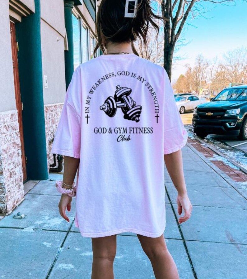 May include: Pink t-shirt with a black graphic of dumbbells and the text "In my weakness, God is my strength. God & Gym Fitness Club."