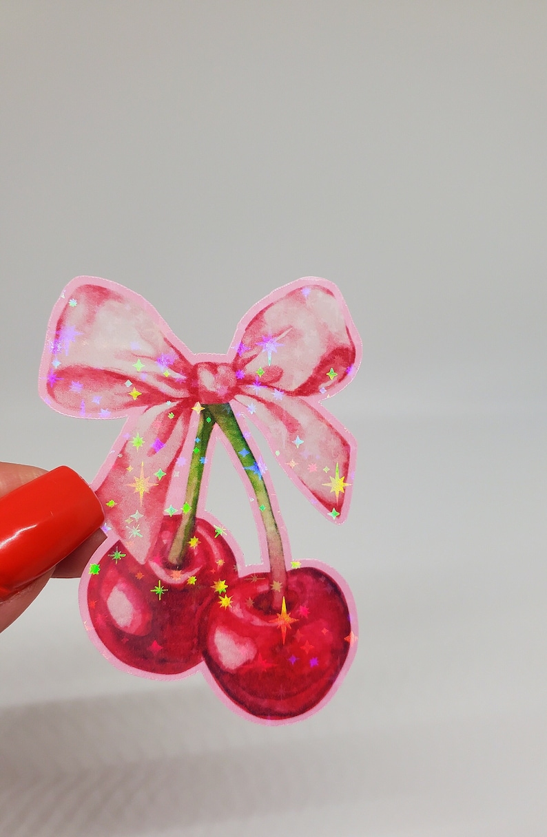 May include: A pink sticker depicting two cherries with a green stalk and a pink bow. The cherries are a deep red colour with sparkling accents. The bow has a watercolour effect.