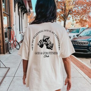 May include: A white t-shirt with a black graphic of dumbbells and the text "In my weakness, God is my strength + God & Gym Fitness Club +" on the back.
