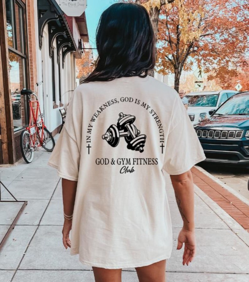 May include: A white t-shirt with a black graphic of dumbbells and the text "In my weakness, God is my strength + God & Gym Fitness Club +" on the back.
