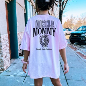May include: Pink oversized t-shirt with black text that reads "Muscle Mommy" and a graphic of a heart with flowers. The text below the graphic reads "Strength - Beauty - Resilience".