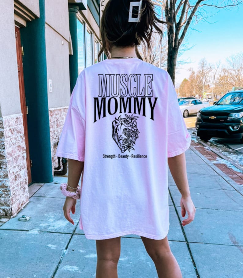 May include: Pink oversized t-shirt with black text that reads "Muscle Mommy" and a graphic of a heart with flowers. The text below the graphic reads "Strength - Beauty - Resilience".