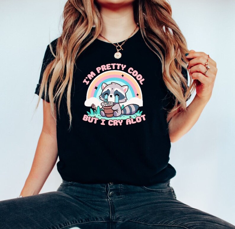 May include: Black t-shirt with a graphic of a crying raccoon holding a cup of coffee. The text on the shirt reads "I'm pretty cool but I cry alot" with a rainbow behind the raccoon.