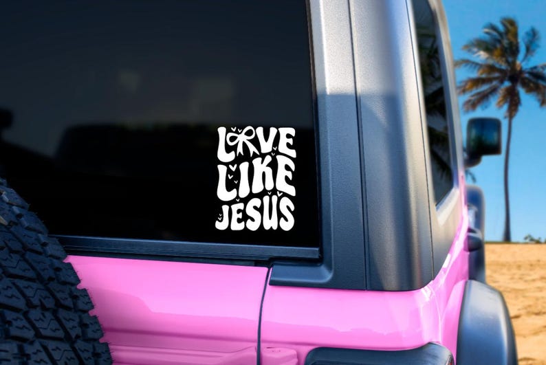 May include: A white vinyl sticker on a pink vehicle window, featuring the words "LOVE LIKE JESUS" in a stylized font. The background shows a palm tree and a blue sky.