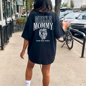 May include: Black oversized t-shirt with white text that reads "Muscle Mommy" and a floral heart graphic. The text below the graphic reads "Strength - Beauty - Resilience".