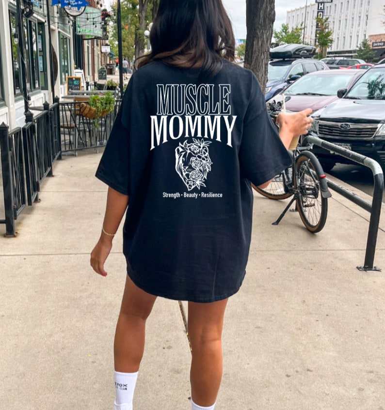 May include: Black oversized t-shirt with white text that reads "Muscle Mommy" and a floral heart graphic. The text below the graphic reads "Strength - Beauty - Resilience".