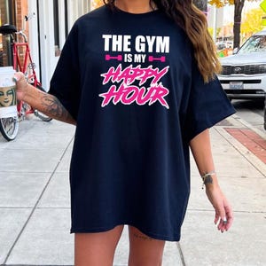 Peut inclure: T-shirt noir surdimensionn&eacute; avec le texte "The Gym is my Happy Hour" en lettres blanches et roses. Le texte est stylis&eacute; avec une halt&egrave;re rose de chaque c&ocirc;t&eacute; de "is my".
