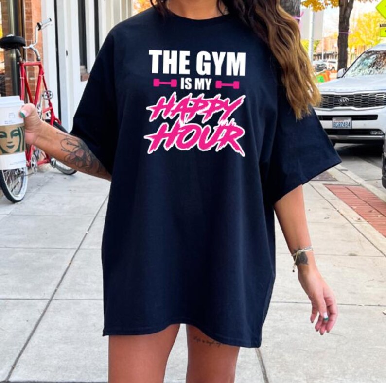Peut inclure: T-shirt noir surdimensionn&eacute; avec le texte "The Gym is my Happy Hour" en lettres blanches et roses. Le texte est stylis&eacute; avec une halt&egrave;re rose de chaque c&ocirc;t&eacute; de "is my".