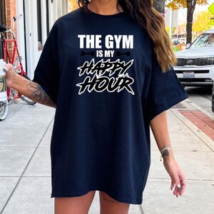 Peut inclure: Un t-shirt noir avec du texte blanc qui dit "The Gym is my Happy Hour" avec un graphique de barre sur chaque c&ocirc;t&eacute; du texte.