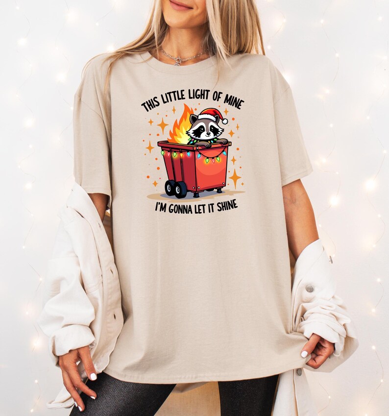 May include: A beige t-shirt with a graphic of a raccoon wearing a Santa hat and sitting in a red trash can with Christmas lights. The text on the shirt reads "This little light of mine I'm gonna let it shine."