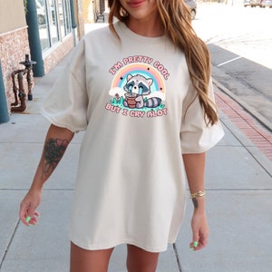 May include: A beige oversized t-shirt with a graphic of a cartoon raccoon crying in a flower pot. The text on the shirt reads "I'm pretty cool but I cry alot".