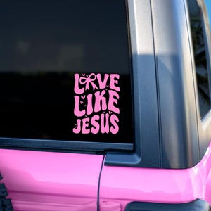 May include: A pink vinyl decal with the words "LOVE LIKE JESUS" in a retro font, applied to the window of a pink vehicle. The decal features a bow and heart accents. The background includes a palm tree and blue sky.