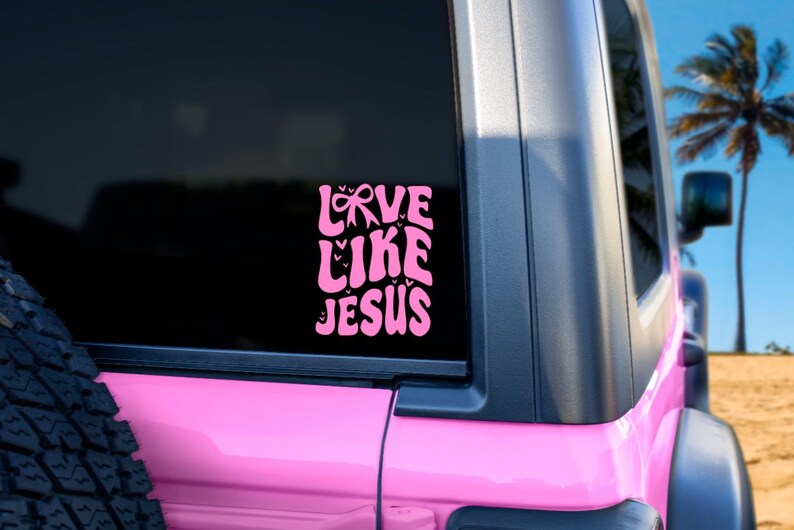 May include: A pink vinyl decal with the words "LOVE LIKE JESUS" in a retro font, applied to the window of a pink vehicle. The decal features a bow and heart accents. The background includes a palm tree and blue sky.