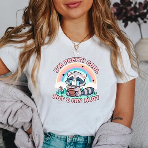 May include: White t-shirt with a graphic of a crying raccoon holding a cup of coffee. The text on the shirt reads "I'm pretty cool but I cry alot".